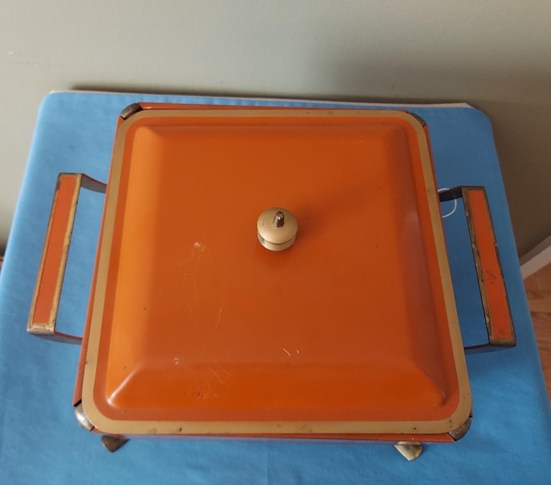 May include: An orange metal serving dish with a square shape and a lid. The dish has a gold trim and handles, and a small, off-white knob on the lid. The dish is sitting on a blue surface.