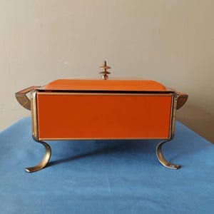 May include: An orange and gold vintage serving dish with a lid. The rectangular dish has a bright orange exterior and gold-colored accents. It stands on four curved gold legs and has a decorative finial on the lid.