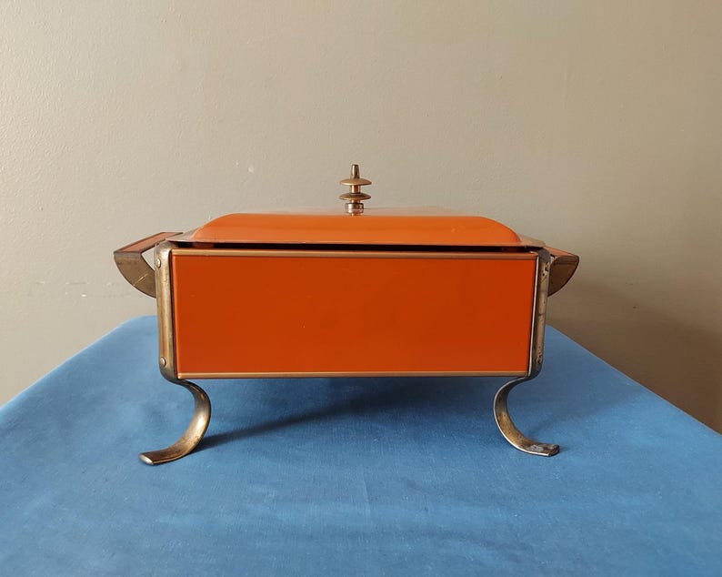 May include: An orange and gold vintage serving dish with a lid. The rectangular dish has a bright orange exterior and gold-colored accents. It stands on four curved gold legs and has a decorative finial on the lid.