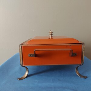 May include: An orange metal box with a handle and decorative feet. The box has a lid with a decorative finial. The box is sitting on a blue surface.
