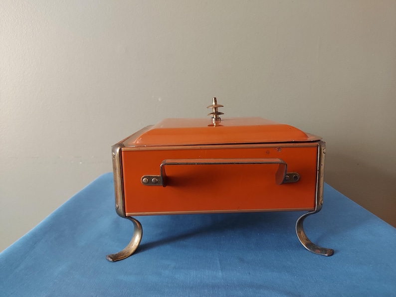 May include: An orange metal box with a handle and decorative feet. The box has a lid with a decorative finial. The box is sitting on a blue surface.