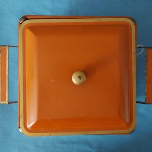 May include: Overhead view of a vintage orange metal serving tray with a square shape and a cream-colored knob. The tray has gold-colored trim and handles. The background is a solid blue color.