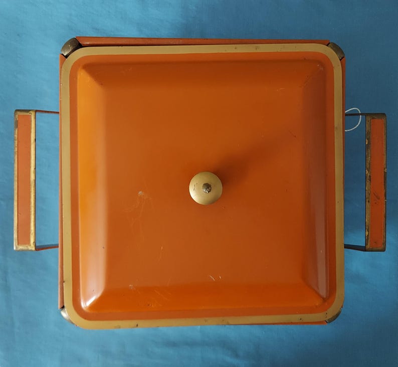 May include: Overhead view of a vintage orange metal serving tray with a square shape and a cream-colored knob. The tray has gold-colored trim and handles. The background is a solid blue color.