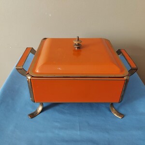 May include: An orange vintage casserole dish with a lid, handles, and decorative feet. The dish has a square shape and a gold-colored trim. The lid is topped with a small, gold-colored knob.
