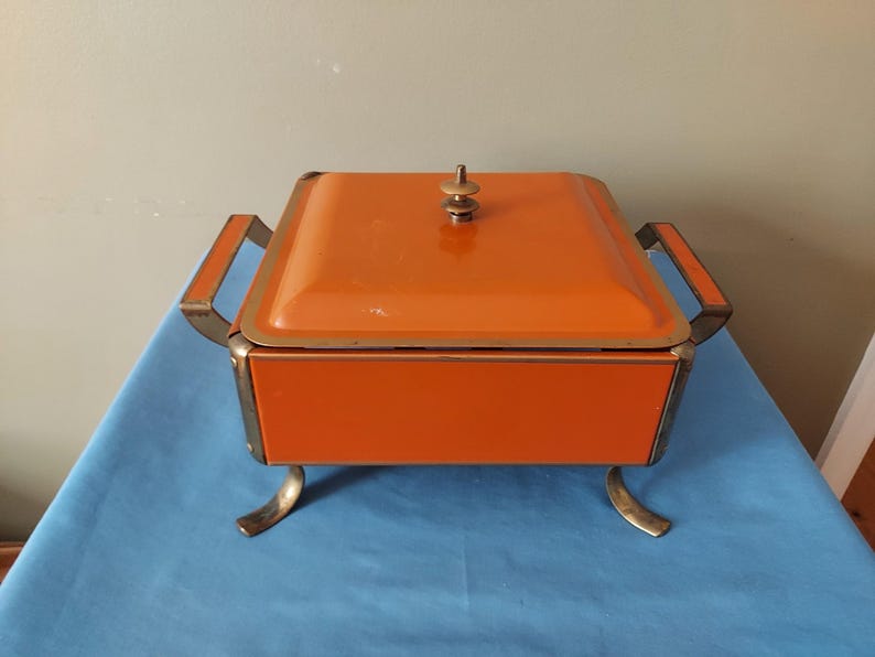 May include: An orange vintage casserole dish with a lid, handles, and decorative feet. The dish has a square shape and a gold-colored trim. The lid is topped with a small, gold-colored knob.
