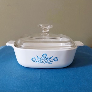 May include: White casserole dish with a clear glass lid. The dish features a blue floral design on the front. The dish has a rectangular shape with rounded corners and two handles. The lid has a knob for easy handling.