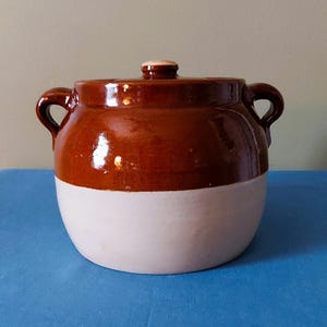 Może przedstawiać: Okrągły ceramiczny garnek z pokrywką, posiadający błyszczącą ciemnobrązową glazurę w górnej części oraz matową jasnobeżową dolną część. Naczynie posiada dwa boczne uchwyty oraz mały uchwyt na środku pokrywki, tworząc dwukolorowy wygląd.