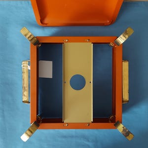 May include: An orange metal box with a matching lid, featuring a square frame with gold-colored legs and handles. The interior has a blue base and a central, vertical, cream-colored panel with a circular opening. A small white tag is attached.