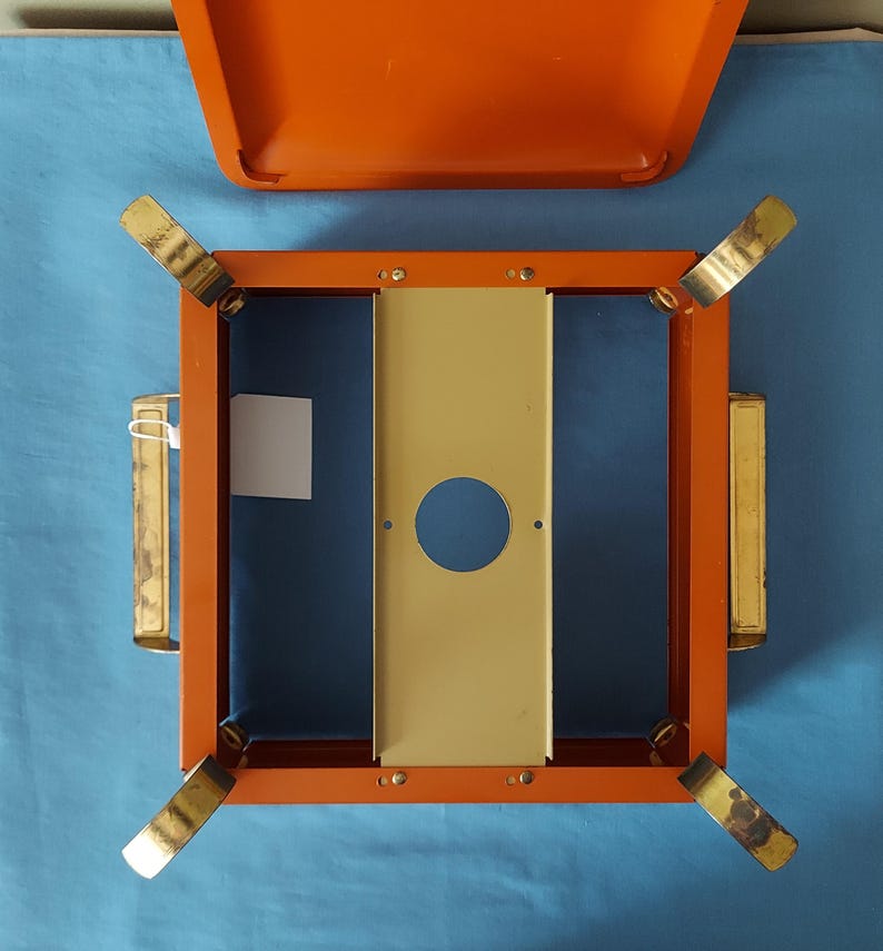 May include: An orange metal box with a matching lid, featuring a square frame with gold-colored legs and handles. The interior has a blue base and a central, vertical, cream-colored panel with a circular opening. A small white tag is attached.