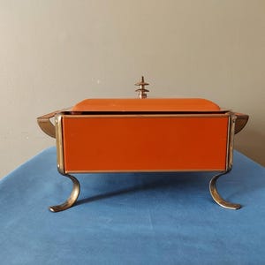 May include: An orange and gold vintage serving dish with a lid. The rectangular dish has curved gold legs and handles. The lid is topped with a decorative gold finial. The dish is set on a blue surface.