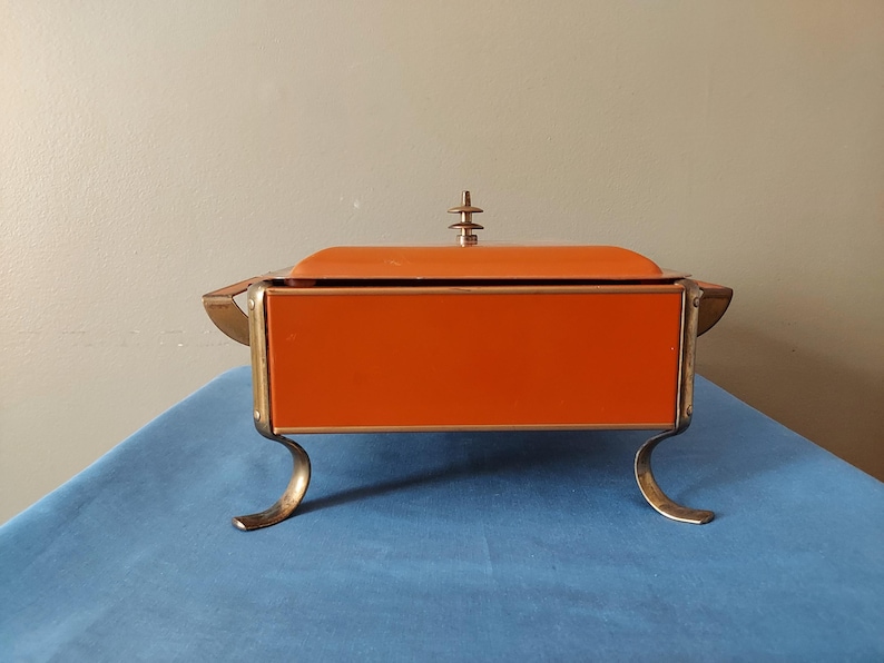 May include: An orange and gold vintage serving dish with a lid. The rectangular dish has curved gold legs and handles. The lid is topped with a decorative gold finial. The dish is set on a blue surface.