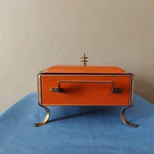 May include: An orange vintage box with a hinged lid, a handle, and decorative gold-colored trim. The box has curved gold-colored feet and a decorative finial on top. The box is displayed on a blue surface.