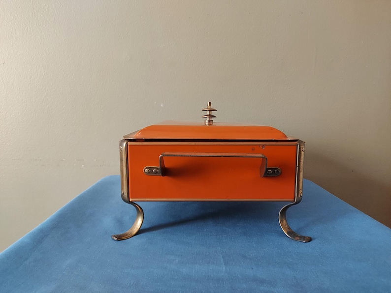 May include: An orange vintage box with a hinged lid, a handle, and decorative gold-colored trim. The box has curved gold-colored feet and a decorative finial on top. The box is displayed on a blue surface.
