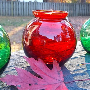 May include: Four vibrant glass vases in green and red hues are displayed on a wooden surface. The vases are spherical with a wide opening. A red leaf lies nearby, adding a seasonal touch. Each vase is approximately 15 cm in diameter.