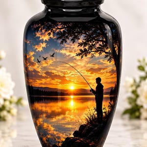 May include: A black metal cremation urn featuring a glossy finish and a wrap-around graphic of a person fishing by a lake at sunset. The scene depicts a silhouette of a person holding a fishing rod, trees, birds, and a bright orange sun.