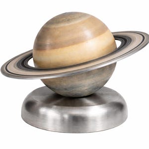 Puede incluir: Escultura decorativa de Saturno. El planeta es de color marrón y beige, con un anillo gris y negro. La escultura se asienta sobre una base de metal plateado.