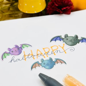 May include: Close-up view of a Halloween-themed craft project.  Three adorable cartoon bats in purple, teal, and gray are arranged on white cardstock with the words 