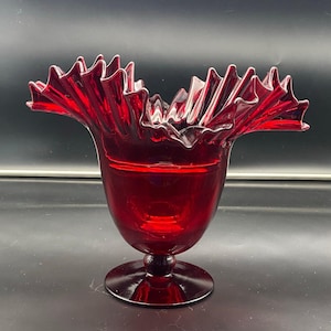 May include: A deep red glass vase featuring a wide, ruffled, and crimped rim. The vessel has a smooth, rounded body that tapers down to a short, thick stem and a circular, flat base. The glass is translucent and uniform in its dark ruby color.
