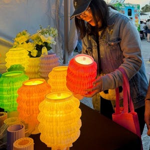 May include: A collection of cylindrical lamps with a pleated, wavy texture in vibrant colors including orange, yellow, and green. The lamps are illuminated from within, casting a warm glow. Some lamps are displayed on a table alongside small vases.