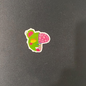 May include: A green and pink cartoon bear sticker with a swirl design on a black background.