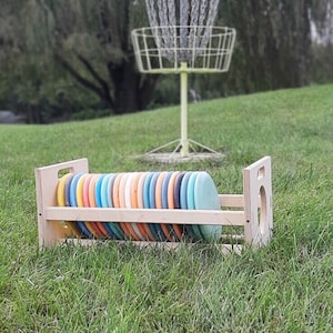 Puede incluir: Un estante de madera con una pila de frisbees de colores. El estante está hecho de madera natural y tiene un diseño sencillo. Los frisbees están dispuestos en una pila ordenada, con los colores alternando entre azul, naranja y amarillo.