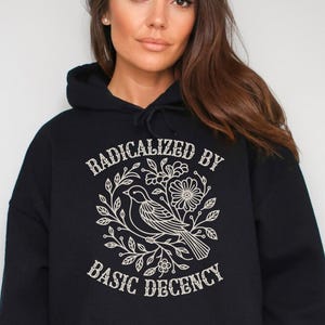 以下が含まれることがあります： 「RADICALIZED BY BASIC DECENCY」の文字が白でプリントされた黒いパーカー。鳥と花の模様がデザインされています。カジュアルな衣服で、パーカーは快適な衣服です。