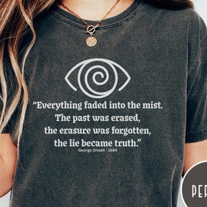 May include: Dark gray t-shirt featuring a white eye graphic and a quote from George Orwell's 1984. The quote reads, "Everything faded into the mist. The past was erased, the erasure was forgotten, the lie became truth." The shirt has a round neck.