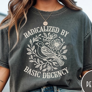 May include: Pepper-colored t-shirt with a white graphic of a bird surrounded by flowers and leaves, with the text "RADICALIZED BY BASIC DECENCY" in a circular design. The shirt has short sleeves and a relaxed fit.