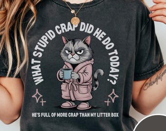 May include: Dark grey t-shirt with a grumpy cat illustration wearing a pink robe and holding a mug. Text includes "WHAT STUPID CRAP DID HE DO TODAY?" and "HE'S FULL OF MORE CRAP THAN MY LITTER BOX."