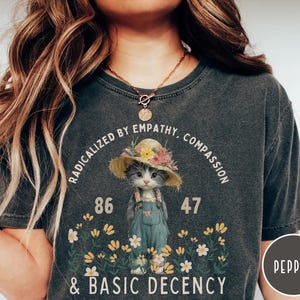 Puede incluir: Una camiseta gris carbón con un gráfico de un gatito con sombrero de paja con flores y peto azul. El texto dice "RADICALIZED BY EMPATHY, COMPASSION & BASIC DECENCY" con los números "86" y "47" y flores de color amarillo y blanco.