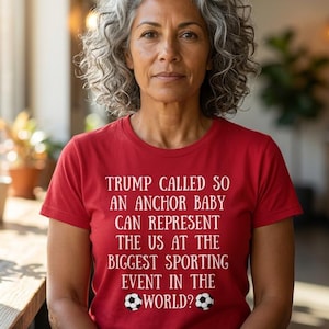 May include: A red short-sleeved t-shirt featuring white text that reads: TRUMP CALLED SO AN ANCHOR BABY CAN REPRESENT THE US AT THE BIGGEST SPORTING EVENT IN THE WORLD? The text is accompanied by two small black and white soccer ball graphics.