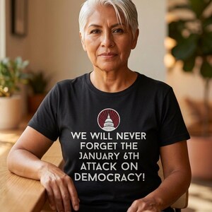 May include: A black short-sleeved t-shirt featuring a white graphic of the U.S. Capitol building dome inside a circle with red rays, and the text: WE WILL NEVER FORGET THE JANUARY 6TH ATTACK ON DEMOCRACY!