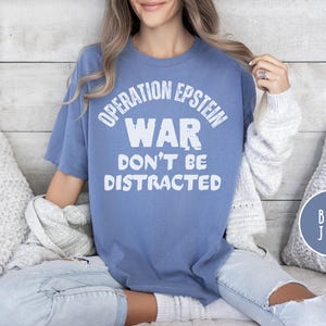 Anti War with Iran Operation Epstein Don't be Distracted. This garment speaks with conviction. A heavyweight, garment-dyed tee printed with a bold, protest-forward message that draws attention without shouting. The soft, broken-in hand of garment dyeing and the relaxed fit make it comfortable for long days at rallies, vigils, or standing on the street holding a sign. Worn by someone who wants their stance to be seen and felt, it layers easily under a jacket or stands alone as a statement piece.