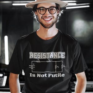 May include: A black t-shirt with a white graphic of a circuit diagram and the text "Resistance Is Not Futile". The person wearing the shirt is smiling and wearing a brown hat and glasses.