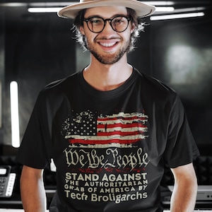 May include: A black t-shirt with a distressed American flag graphic and the text "We the People Stand Against the Authoritarian Grab of America by Tech Broligarchs".