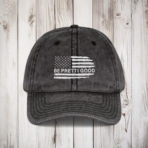 May include: A dark grey baseball cap with a curved peak. The cap displays a distressed American flag graphic and the text "BE PRETTI GOOD" in white. The hat has a traditional six-panel construction.