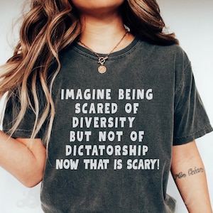 May include: Dark gray t-shirt with the text "IMAGINE BEING SCARED OF DIVERSITY BUT NOT OF DICTATORSHIP NOW THAT IS SCARY!" in white. The shirt has a crew neck and short sleeves. A gold necklace is visible.