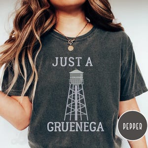 May include: A dark gray t-shirt with the text "Just a Gruenega" and a graphic of a water tower with the text "Gruene" on it. The word "Pepper" is visible in a circle in the lower right corner of the image.