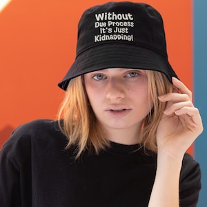 May include: Black bucket hat with the text "Without Due Process It's Just Kidnapping!" in white. The hat is worn by a person wearing a black top.