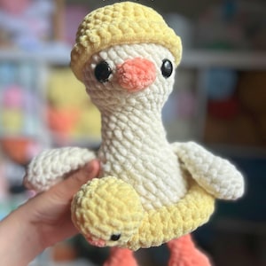 May include: A handmade crochet goose toy with a yellow hat and floaty. The goose has a white body, black eyes, and an orange beak and feet. The text "GOOSE FLOATY PATTERN (PDF ONLY)" is at the top of the image.