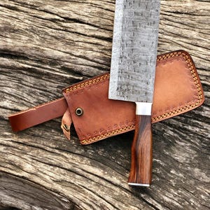 May include: A Damascus steel cleaver with a wooden handle and a leather sheath. The cleaver has a rectangular blade with a patterned design. The handle is dark brown and the sheath is light brown with decorative stitching.