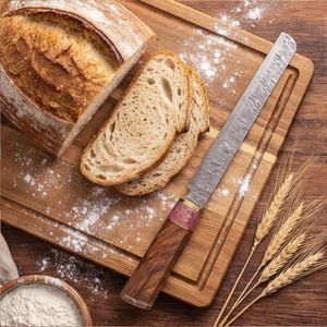 May include: A loaf of sliced bread and a bread knife rest on a wooden cutting board. The bread knife has a patterned blade and a wooden handle with a pink accent. Wheat stalks and flour are also visible.