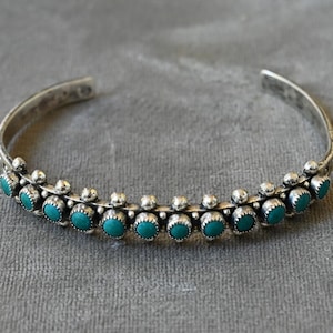 May include: A silver cuff bracelet featuring a row of small, round, vibrant turquoise stones set in scalloped bezels, accented by polished silver spheres between each stone, creating a textured, metallic band with an open back design.