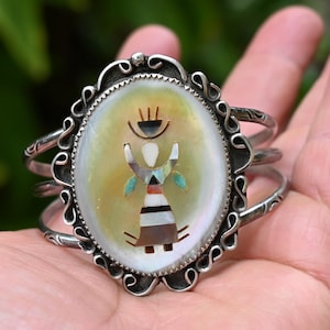 May include: A silver cuff bracelet featuring an oval mother-of-pearl inlay with a central mosaic figure. The figure is composed of small pieces of shell and stone in white, black, and turquoise. The silver frame has a decorative scalloped edge.