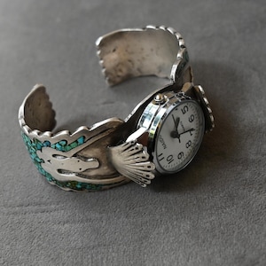 Könnte beinhalten: Eine silberne Manschettenuhr mit weißem Zifferblatt, schwarzen Ziffern und Zeigern. Das Armband ist mit eingelegten Türkis-Steinfragmenten und einem zentralen Vogelmotiv verziert und weist eine texturierte Metalloberfläche an den Rändern auf.