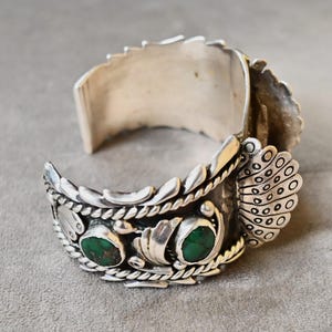 May include: A silver cuff bracelet featuring two oval green turquoise stones set in metal. The band is decorated with twisted rope wire, leaf-like metal accents, and a fan-shaped element with circular cutouts on the side of the cuff.