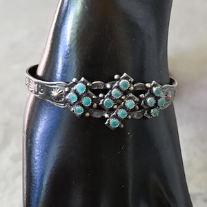 May include: A silver cuff bracelet featuring a central cluster of small, round, light blue turquoise stones arranged in a geometric pattern. The metal band is oxidised and decorated with stamped sunburst and floral motifs along the sides.