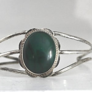 May include: Silver cuff bracelet with a large, oval, dark green stone. The stone is set in a decorative bezel with a rope-like border. The bracelet has a simple, open design with three parallel bands.