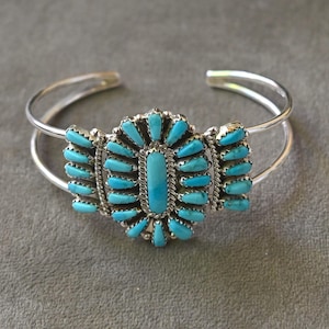 May include: A silver cuff bracelet featuring a central cluster of small, oval-shaped turquoise stones arranged in a radial pattern. The metal band is split into two thin, polished silver wires that connect to the decorative stone setting.