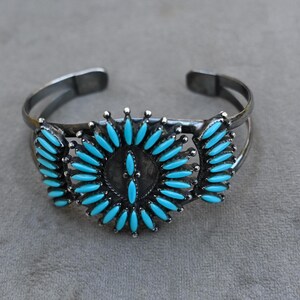 Op de afbeelding: Zilverkleurige manchetarmband met turquoisekleurige steenaccenten. De armband heeft een centraal cirkelvormig ontwerp en twee zijontwerpen, allemaal versierd met de helderblauwe stenen. De armband is aan de achterkant open.